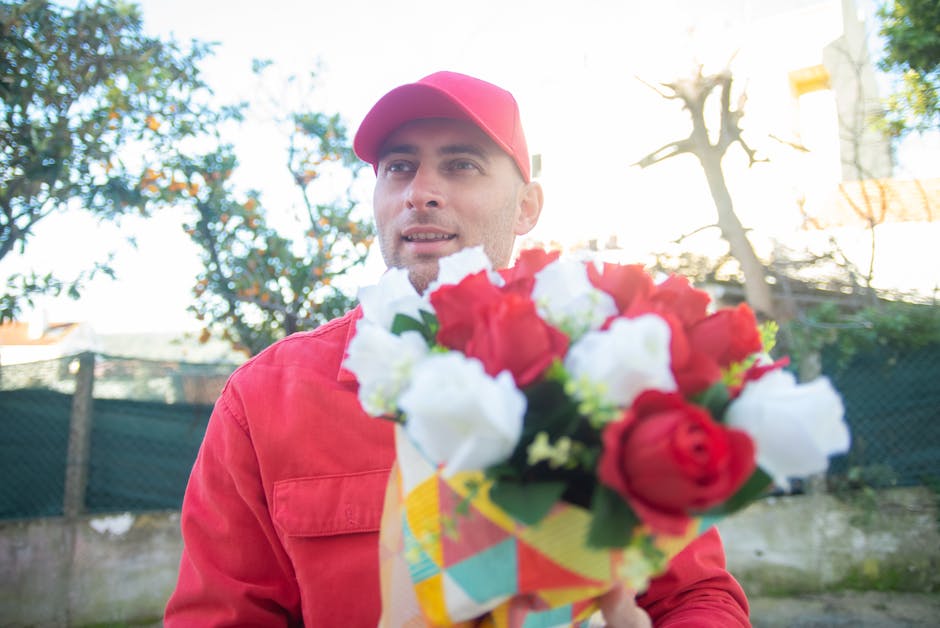 A delivery person wearing a red cap and red jacket holds out a vibrant floral bouquet towards the camera. The bouquet features red and white roses, with some green foliage, wrapped in colorful paper.