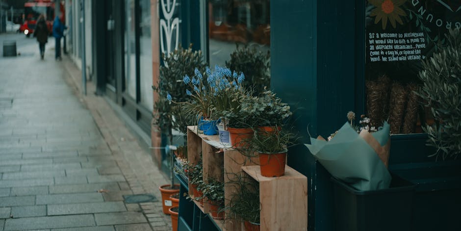 A row of potted flowers arranged on wooden shelves outside a flower shop along a sidewalk. The plants include a mix of greenery and small flowering plants with blue blooms, housed in various terracott