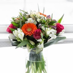 A masterful display of colour and form, this bouquet is a true reflection of Chingford's harmonious landscapes. Creamy-peach and bold orange gerberas create a radiant halo, their petals glowing in a sunlit room reminiscent of Chingford Green. Deep fuchsia roses infuse the centre with luxurious intensity, while ivory alstroemeria and small white flowers trace airy patterns between lush, pointed greenery. Aspidistra leaves are artfully wrapped around the stems, grounding the exuberant blooms in tranquil green, just as the River Ching winds through the local parks. Arranged in a tall, clear vase with visible stems and dense foliage, the composition feels fresh and organic, echoing the local commitment to quality and care. Soft morning light enhances the natural beauty of each blossom, evoking a sense of optimism and joyful anticipation. Whether destined for a celebratory brunch, a new home in Highams Park, or a heartfelt gesture for a neighbour, this bouquet sings with the grace and warmth found in every Chingford gathering.